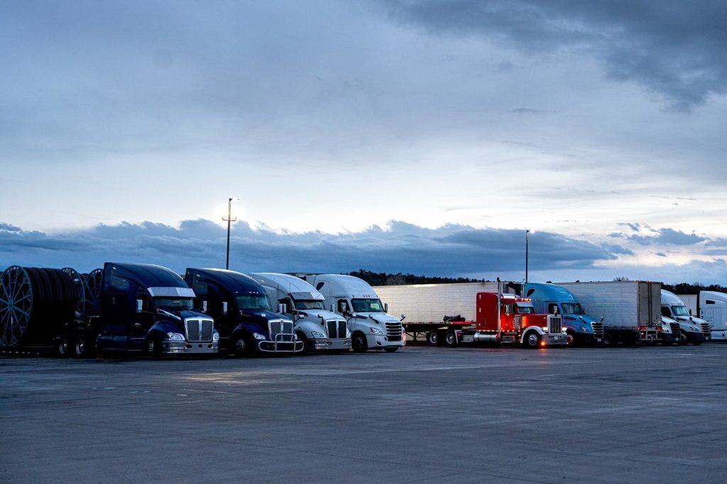 A row of parked semi-trucks in a lot during dusk, with a cloudy sky in the background.